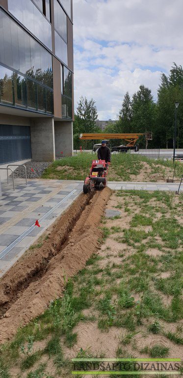 Darbuotojas tranšėjakase kasa siaurą tranšėją šalia pėsčiųjų tako