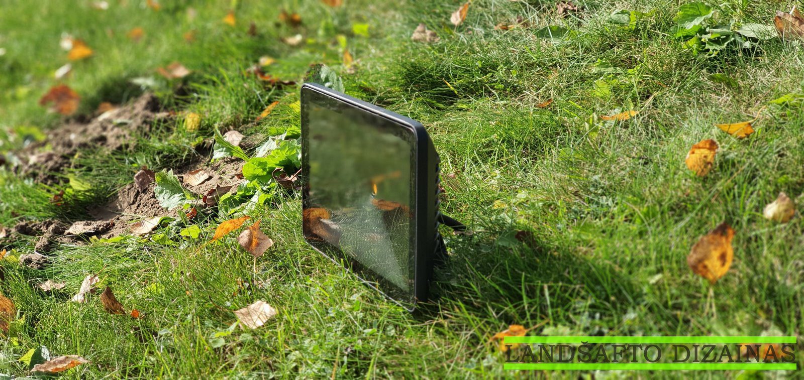 Juodas LED prožektorius pastatytas žalioje vejoje tarp nukritusių lapų