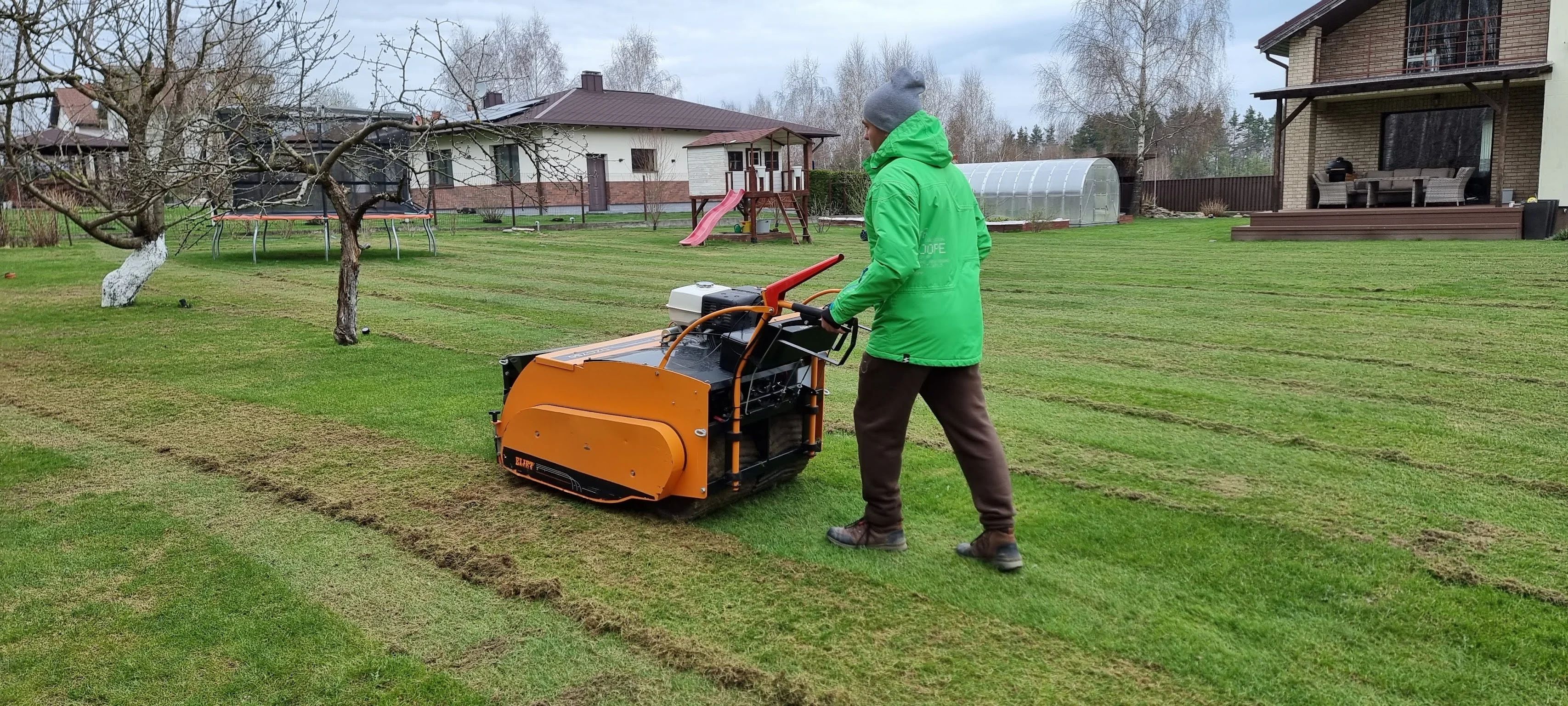Darbuotojas su oranžine „Eliet“ atsėjimo mašina atnaujina veją namo kieme
