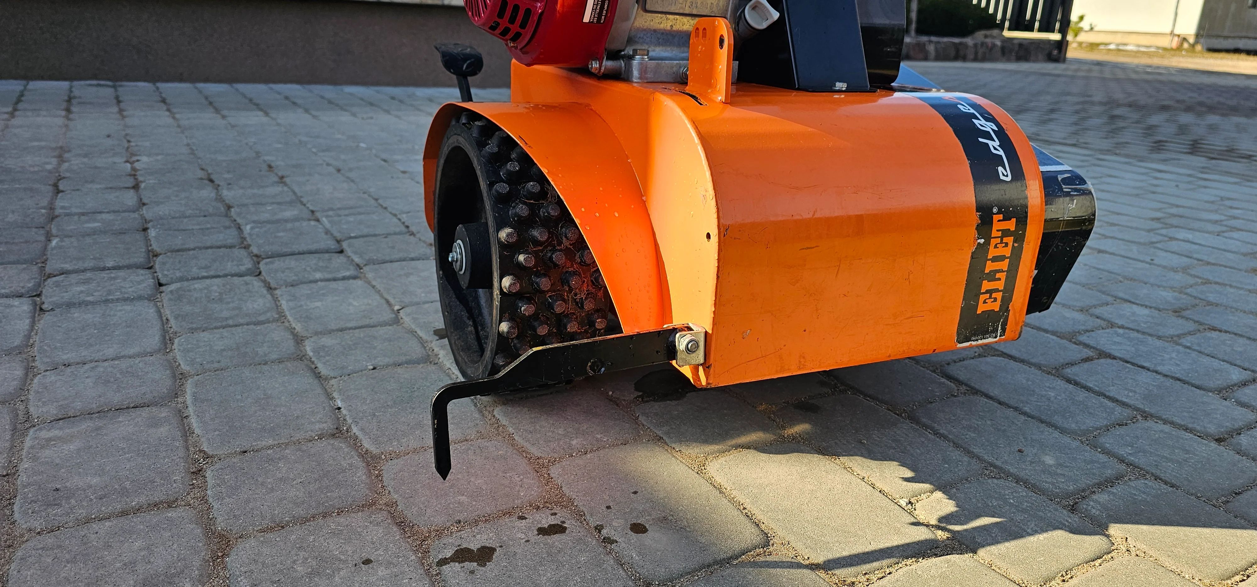 Oranžinė vejos kraštų pjovimo mašina ELIET su dygliuotu ratu ant grindinio trinkelių
